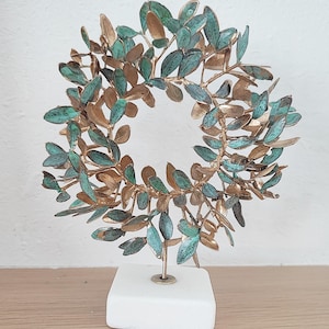 May include: A circular sculpture featuring metallic gold and oxidised teal green leaves arranged in a wreath pattern. The foliage is attached to a thin vertical metal rod that is securely mounted onto a solid, square, white stone base.