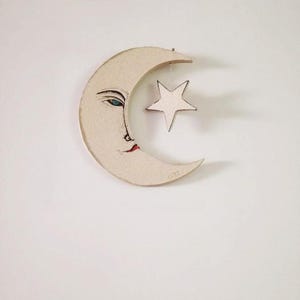 Puede incluir: Una luna creciente blanca con una cara pintada y una pequeña estrella blanca colgando de ella. La luna está hecha de madera y tiene un aspecto rústico y hecho a mano.
