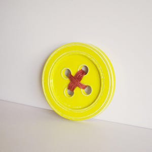 May include: A large, yellow button with four holes and red thread sewn through the holes in a criss-cross pattern.