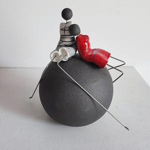 Może przedstawiać: Ceramiczna rzeźba przedstawiająca dwie postacie siedzące na czarnej kuli. Postacie są stylizowane i mają proste rysy. Jedna postać jest ubrana w biało-czarną sukienkę, a druga w czerwoną sukienkę. Rzeźba jest wykonana z gliny i ma rustykalny, ręcznie robiony wygląd.