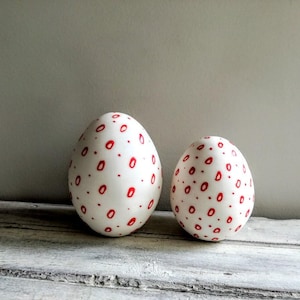 May include: Two white ceramic eggs decorated with red polka dots. The eggs are sitting on a whitewashed wooden surface.