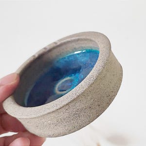 以下が含まれることがあります： テクスチャのあるグレーの外側と内側に青い釉薬をかけた、小さな丸いセラミック製の皿。皿はわずかに盛り上がった縁をしています。