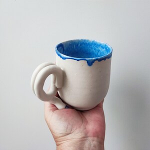 Op de afbeelding: Witte keramische mok met een blauwe rand en een unieke, gedraaide handgreep. De mok wordt in een hand gehouden.