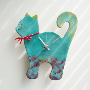 Op de afbeelding: Een turquoise blauwe keramische kattenklok met een rode strik en zilveren wijzers. De klok heeft een verweerde afwerking met hints van goud en rood.