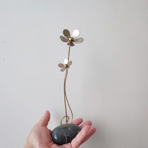 Könnte beinhalten: Eine Metallskulptur aus zwei stilisierten Blumen auf einem Drahtstamm, montiert auf einem runden grauen Steinsockel.