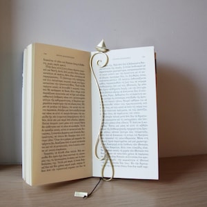 May include: A gold metal bookmark shaped like a sailboat with a curled tail. The bookmark is in a book with white pages.