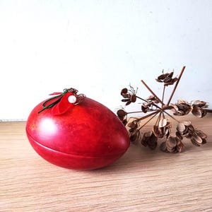 May include: A red egg-shaped trinket box with a green and red floral design on top. The box is sitting on a wooden surface next to a dried flower.