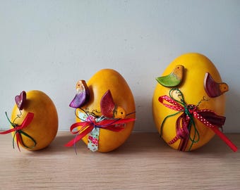 May include: Three yellow Easter eggs decorated with colorful wooden birds and red and white ribbon.