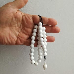 Puede incluir: Un collar de cuentas blancas con un dije de perla blanca. El collar está hecho de cuentas redondas blancas y está enhebrado en un cordón negro.