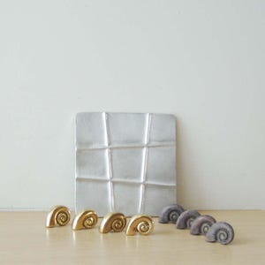 May include: A set of decorative gold and gray snail-shaped ceramic pieces on a white textured square tile.