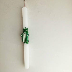 May include: A white candle with a green and white striped ribbon tied around it. The ribbon has a small green shirt design on it.
