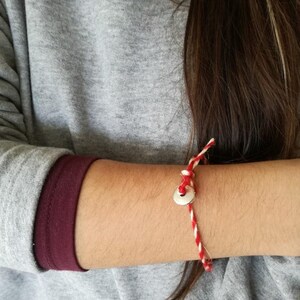 Op de afbeelding: Een rood en wit touwtjesarmband met een zilveren bedel. De armband is om de pols van een persoon gebonden.