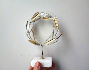 May include: A silver and gold olive branch wreath sculpture on a white marble base.
