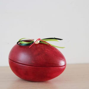 Può includere: Una scatola decorativa rossa a forma di uovo con coperchio, ornata da nastri colorati e una perla. La scatola è realizzata in un materiale liscio e poggia su una superficie di legno chiaro. I nastri sono verdi, neri e rossi.