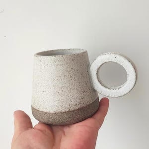 Peut inclure: Une tasse en céramique mouchetée avec un corps arrondi et une anse circulaire. La tasse a une palette de couleurs blanche et marron, avec une base marron plus foncée. La poignée est blanche.