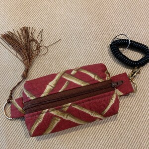 May include: A small red zippered pouch with a brown zipper and a gold key ring. The pouch has a pattern of brown bamboo stalks on a red background. A brown tassel is attached to the pouch.
