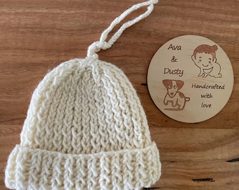 May include: A white knitted baby hat with a tie string. A wooden sign with the text "Ava & Dusty Handcrafted with love" is in the background.