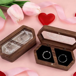 以下が含まれることがあります： 透明な蓋が付いた2つの木製リングボックス。1つは開いており、もう1つは閉じており、銀色のリングが表示されています。ボックスは、花、赤いハート、リボンが置かれたピンク色の表面にあります。閉じたボックスには「Timothy & Florence」という文字があり、開いたボックスには「Milson & Woren」という文字があります。