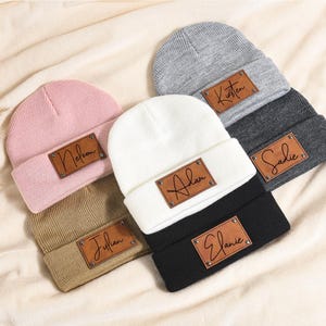 May include: A collection of personalized beanies in various colors, including pink, white, gray, black, and tan. Each beanie features a brown leather patch with a name in a handwritten style font. The beanies are displayed on a cream-colored fabric.