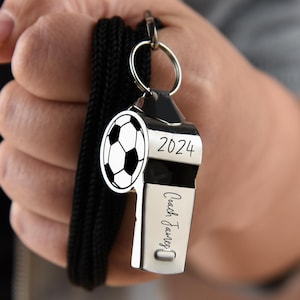 以下が含まれることがあります： 黒い紐が付いた銀色の笛とサッカーボールのチャーム。笛には「2024」の年号と「Coach James」の名前が刻まれています。サッカーボールのチャームは白と黒です。