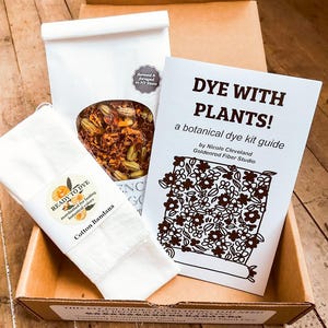 May include: A botanical dye kit in a cardboard box containing a white cotton bandana with a Ready to Dye label, a white pouch of dried orange and yellow plant materials, and a white instructional booklet titled Dye With Plants!