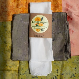 May include: A white silk scarf wrapped in a brown paper band labeled READY TO DYE mordanted for lasting botanical colors Silk Scarf, positioned over a collection of folded fabric squares in shades of orange, pink, yellow, and dark gray.