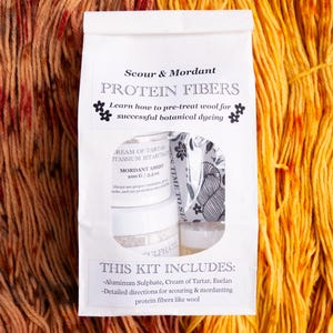 May include: A white paper bag with a clear window, containing a protein fiber kit for botanical dyeing. The kit includes aluminum sulfate, cream of tartar, and detailed instructions. The background features dyed wool in shades of orange and yellow.