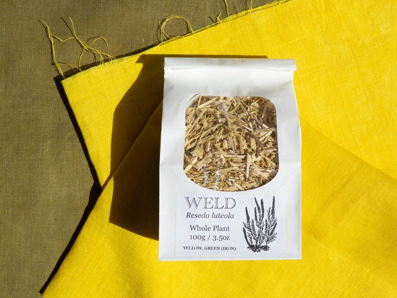 May include: A white paper bag with a clear window containing dried Weld Reseda luteola whole plant material. The label reads Weld, Reseda luteola, Whole Plant, 100g / 3.5oz, YELLOW, GREEN (IRON) and features a black botanical illustration.