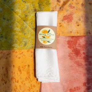 May include: A folded white silk bandana wrapped in a brown paper band with the text READY TO DYE mordanted for lasting botanical colors Silk Bandana, placed over four squares of fabric dyed in yellow, orange, peach, and reddish-pink botanical patterns.