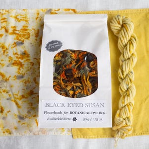 May include: A white paper bag containing dried Black Eyed Susan flowerheads for botanical dyeing, labeled 50 g / 1.75 oz, flanked by a piece of white fabric with yellow and grey mottled patterns and a skein of yellow yarn with grey flecks.