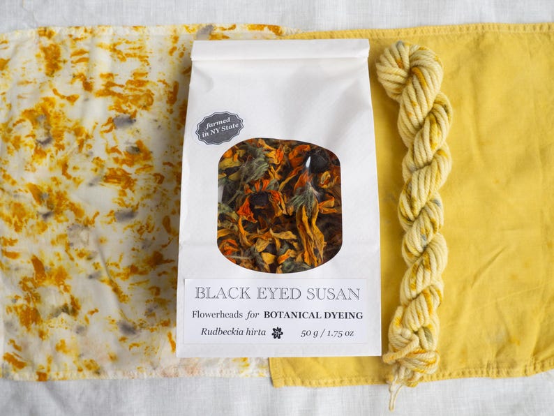May include: A white paper bag containing dried Black Eyed Susan flowerheads for botanical dyeing, labeled 50 g / 1.75 oz, flanked by a piece of white fabric with yellow and grey mottled patterns and a skein of yellow yarn with grey flecks.
