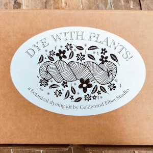 May include: A rectangular brown cardboard box featuring a large oval white sticker on the front. The sticker displays the text DYE WITH PLANTS! and a botanical dyeing kit by Goldenrod Fiber Studio, accompanied by a graphic of yarn and flowers.