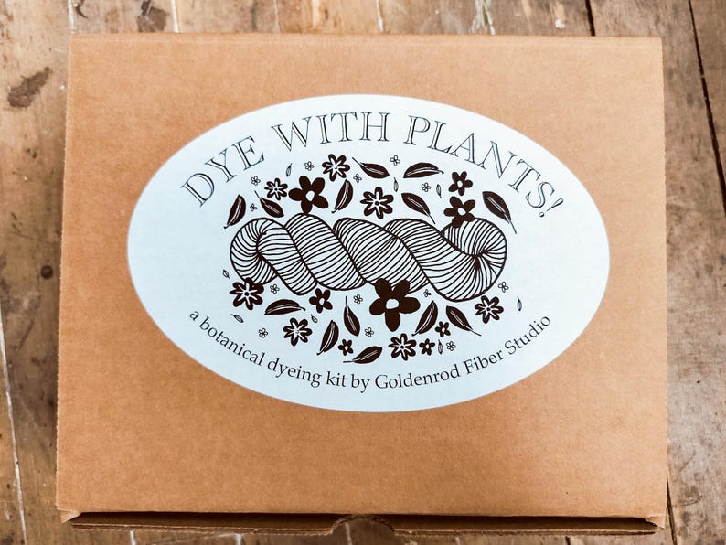 May include: A rectangular brown cardboard box featuring a large oval white sticker on the front. The sticker displays the text DYE WITH PLANTS! and a botanical dyeing kit by Goldenrod Fiber Studio, accompanied by a graphic of yarn and flowers.