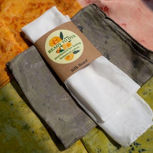 May include: A white silk scarf wrapped in a brown paper band labeled READY TO DYE, mordanted for lasting botanical colors, resting on top of three folded fabric pieces in shades of orange, muted green, and light pink with mottled patterns.