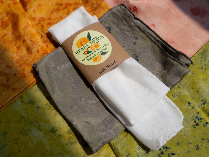 May include: A white silk scarf wrapped in a brown paper band labeled READY TO DYE, mordanted for lasting botanical colors, resting on top of three folded fabric pieces in shades of orange, muted green, and light pink with mottled patterns.