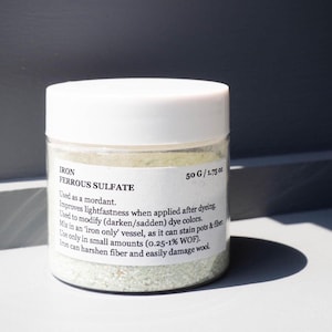 May include: A clear jar with a white lid containing ferrous sulfate, a mordant for dyeing. The label reads "IRON FERROUS SULFATE 50 G / 1.75 oz." The jar is on a white surface with a dark shadow.