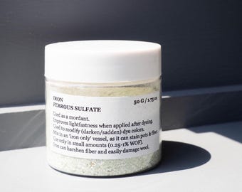 May include: A clear jar with a white lid containing ferrous sulfate, a mordant for dyeing. The label reads "IRON FERROUS SULFATE 50 G / 1.75 oz." The jar is on a white surface with a dark shadow.