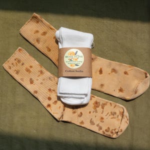 May include: A pair of light tan ribbed cotton socks featuring a scattered brown botanical print, bundled together with a white pair of socks by a brown paper label that reads READY TO DYE mordanted for lasting botanical colors Cotton Socks.