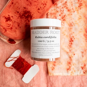 May include: A white plastic jar labeled MADDER ROOT Rubia cordifolia 100 G / 3.5 oz, surrounded by a pair of coral-colored socks with red speckles, a piece of fabric with red mottled patterns, and a white card wrapped with vibrant red thread.