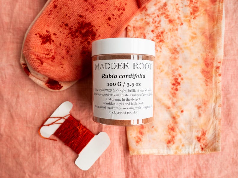May include: A white plastic jar labeled MADDER ROOT Rubia cordifolia 100 G / 3.5 oz, surrounded by a pair of coral-colored socks with red speckles, a piece of fabric with red mottled patterns, and a white card wrapped with vibrant red thread.