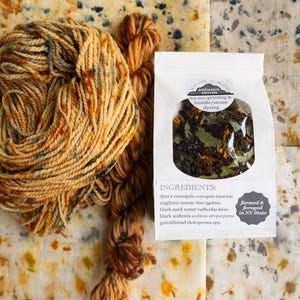 May include: A skein of variegated orange and brown yarn next to a white paper pouch labeled Botanical Blend Autumn Storm for eco-printing and bundle/steam dyeing, containing dried plant matter with a list of ingredients and farmed and foraged in NY State.