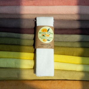 May include: A stack of folded linen fabric in various shades of pink, brown, green, and yellow. A white piece of linen cloth is wrapped in a brown paper band labeled READY TO DYE, mordanted for lasting botanical colors, Linen Cloth, 1/2 Yd. ~28" x 36".