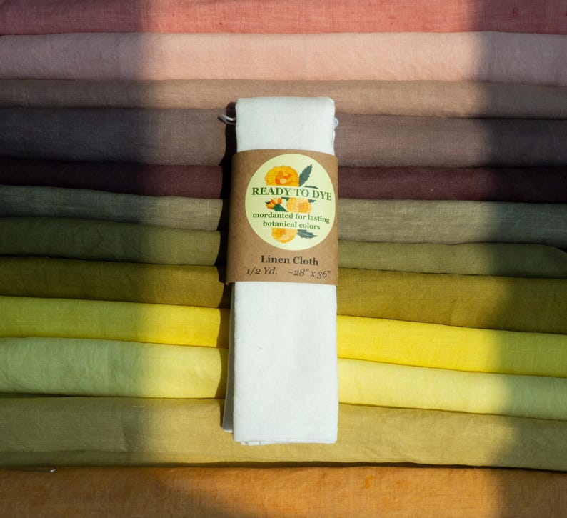May include: A stack of folded linen fabric in various shades of pink, brown, green, and yellow. A white piece of linen cloth is wrapped in a brown paper band labeled READY TO DYE, mordanted for lasting botanical colors, Linen Cloth, 1/2 Yd. ~28" x 36".