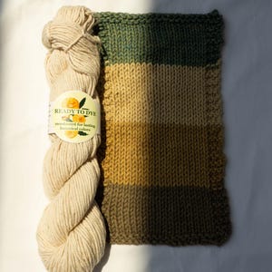 May include: A skein of off-white yarn with a label reading READY TO DYE mordanted for lasting botanical colors, positioned next to a rectangular knitted swatch featuring horizontal stripes in shades of dark green, tan, and mustard yellow.