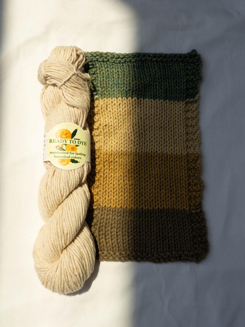 May include: A skein of off-white yarn with a label reading READY TO DYE mordanted for lasting botanical colors, positioned next to a rectangular knitted swatch featuring horizontal stripes in shades of dark green, tan, and mustard yellow.