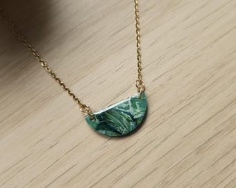 Puede incluir: Un collar de cadena dorada con un colgante. El colgante tiene forma de media luna con un diseño veteado verde y blanco. El collar se exhibe sobre una superficie de madera marrón claro.