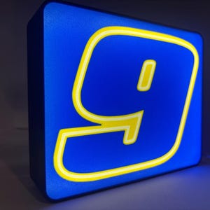 May include: A rectangular LED light box sign featuring a vibrant blue background with a large, stylized yellow number nine centered on the front face. The illuminated design has a bold, modern aesthetic with clean lines and a glowing neon-like appearance.