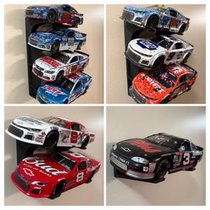Könnte beinhalten: Eine Sammlung von Miniatur-NASCAR-Rennwagen in verschiedenen Farben und Designs, die auf schwarzen, an der Wand befestigten Regalen ausgestellt sind. Die Autos verfügen über Sponsorenlogos und Rennnummern, die eine Vielzahl von Marken und Stilen zeigen. Die Sammlung umfasst Autos mit den Nummern 8, 88 und 9.