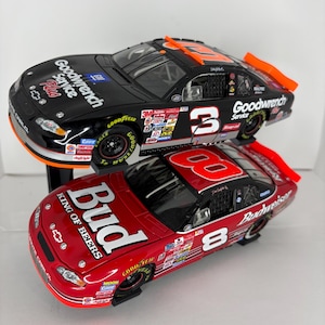 Pode incluir: Dois modelos de carros de corrida em miniatura empilhados verticalmente. O carro de cima é preto com o número 3 e a marca Goodwrench. O carro de baixo é vermelho com o número 8 e a marca Budweiser. Ambos os modelos Chevrolet Monte Carlo têm decalques.
