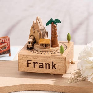 May include: Wooden music box featuring a mountain, palm tree, house, and train design. The box is personalised with the name "Frank".